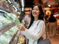 Asiatische Frau steht lächelnd an einem Supermarktregal für Gemüse